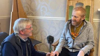 Tudur Hallam yn eistedd yng nghadair yr Eisteddfod a Dei Tomos yn ei holi 