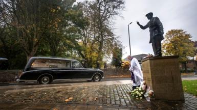 A hearse passing a statue with mourners lining the route