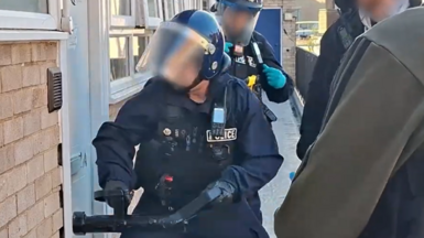 Police officer in tactical gear breaking a door down using a battering ram tool. There are other officers nearby. Their faces have been blurred,