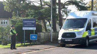 A female police officer stands at the entrance to Gainsborough Care Home. A marked police van is parked on the roadside. The two-storey building can just be seen behind some trees and a blue sign saying 'Gainsborough Care Home'.