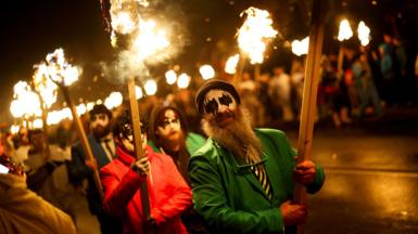 Participants carry fire sticks while wearing masks andf hats and colourful suits during the procession at night.
