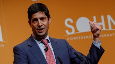 Kevin Warsh wearing a blue suit, light blue shirt and red patterned tie speaking at a conference in front of an orange background