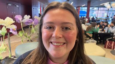 A close-up image of Amy Smith. She is a young woman with long, brown hair and she is smiling at the camera. She is sitting in front of some fake potted flowers and behind her are students sitting at tables.