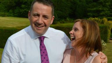 John Hunt is in a white shirt and wearing a purple tie with black spots. His wife Carol is facing him and laughing. They're standing outside on grass.