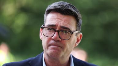 Prime Minister Andy Burnham wears dark-rimmed glasses and a blue suit.