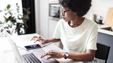 Woman using laptop, notepad, and calculator to look at bills on a white kitchen table