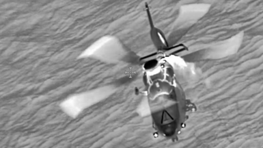 A black and white photo of a helicopter over the sea with rotors turning.