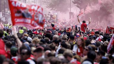 A dense crowd of Arsenal football supporters celebrates in a city street filled with red smoke. In the foreground, a large red banner is blurred, while in the mid-ground, a fan stands above the crowd with arms raised, holding a smoking flare.