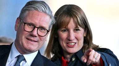 Sir Keir Starmer and Rachel Reeves with their heads together look towards the camera. Reeves is pointing with one hand