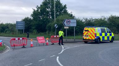 A police officer stands next to a high-visibility police van at a road closure checkpoint blocking a junction.Red and white plastic barriers, orange traffic cones, and a red "ROAD CLOSED" sign block the left lane.Behind the officer, a white directional road sign points right toward Ampthill (2¾ miles) and features a 7.5-tonne weight limit restriction. A smaller sign below it points left toward Houghton Fields.The background is filled with dense green trees and a cloudy, overcast sky.