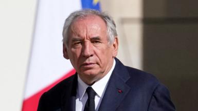 Prime Minister François Bayrou dressed formally in a dark suit, white shirt, and dark tie. He has grey hair and serious expression on his face. He’s standing outdoors, and behind him are two partially visible flags — one appears to be the French tricolor (red, white, and blue), and the other is the European Union flag. There is a small red pin on his left lape