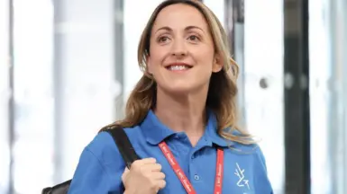 Natalie Cassidy stands in a building, smiling, wearing the blue polo shirt of North Hertfordshire College and carrying a black rucksack