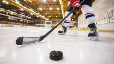 An image of an unseen hockey player wearing a red glove and striped white, red and blue socks hitting a puck along the ice
