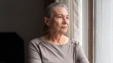 An older woman with long silver hair looks out a bedroom window. She wears a pale grey top with silver beads around the shoulders.
