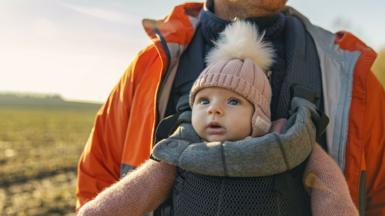 A smiling baby wearing a pink hat is looking out while being carried in a grey baby sling worn by her father, who wears a thick blue jumper and orange rain jacket.