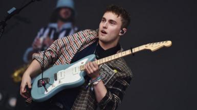 Sam Fender in a chequered open shirt with light blue guitar on stage.