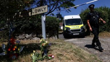 A police officer is seen walking beneath a cordon with a police van in the background and to the left a Drive Slowly sign with flowers underneath it.