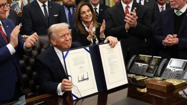 US President Donald Trump signs document to end shutdown, holding it up at his desk which a crowd of people around him applaud.