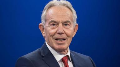 Tony Blair, wearing a red tie and blue suit jacket, and set against a plain blue background, looks directly at the camera