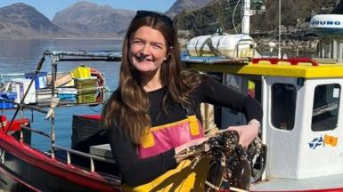 Brooke Lamond holds a lobster in front of a small creel fishing boat. She wears a long-sleeve black top with yellow fishing overalls on top. It is a sunny day and you can see the water behind her and hills in the distance.