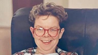 A photo from the 80s of a woman in a leather arm chair wearing round red glasses and smiling