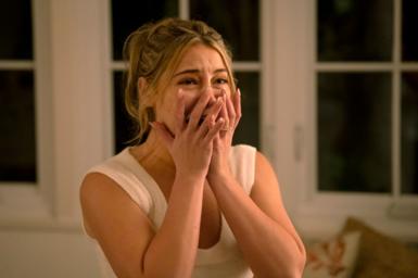 A still from I Know What You Did Last Summer shows actress Madelyn Cline with her hands clasped to her face, mid-scream. She's inside a house at night with large bay windows behind her.