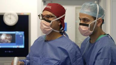 Two male surgeons in blue scrubs and face masks operate on a patient in a hospital. One of the surgeons is holding a white instrument in his hand. Behind them a screen in showing what they are operating on, but this is blurred. 