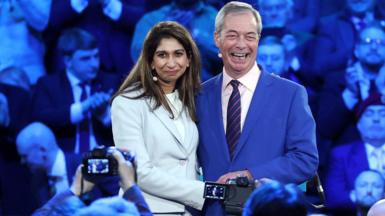Suella Braverman and Nigel Farage with arms around each other smiling for cameras in front of a crowd. 