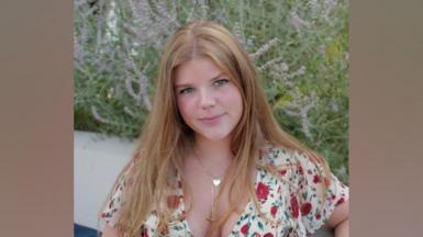 Orla Wates, a girl with long, ginger hair, smiles at camera. She is wearing a white top with red flowers and sits in front of a flowering lavender plant.
