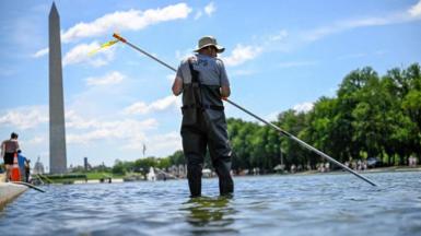A worker in waders seen using a long pole to clean the pool