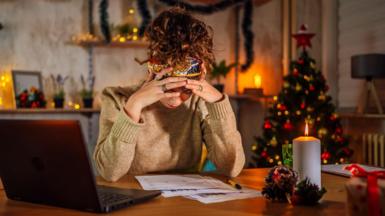 woman in beige jumper in front of a computer with her head in her hands. Xmas tree in the background
