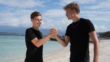 Alfie Watts and Owen Wood smiling and doing a bro shake on a beach in a hot country. Both are wearing black, with clear waters and white sand visible behind them