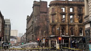 The fire-ruined blackened remains of a sandstone building on the corner of a junction in Glasgow city centre. The scene is cordoned off with yellow tape and the building's rubble surrounds the building perimeter.