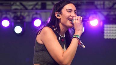 Julia Wolf sings on stage in a grey tank top and beaded necklace in Austin, Texas. She has dark hair.