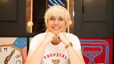 Maisie Adam poses, smiling, resting her chin on her knuckles. She's wearing a white t-shirt with the word "tropical" in a red font above a hand-drawn picture of a bunch of bananas.
