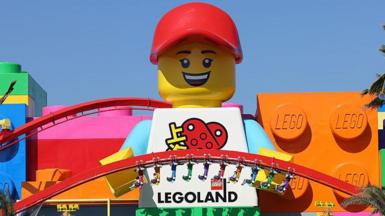 A photo of a towering Lego minifigure mascot standing between two red rollercoaster tracks in Legoland Shanghai. 