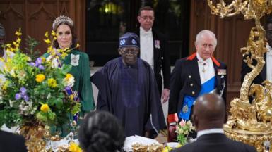 The Princess of Wales in a green dress President of Nigeria Bola Ahmed Tinubu in navy blue and King Charles III in a black suit stand at the table which is decorated with gold features and spring flowers