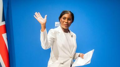 Kemi Badenoch wearing a white jacket and white t-shirt is pictured waving to the camera. There is a union jack pictured to the left.