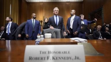 Robert F Kennedy Jr stands behind a chair, pushed up to a wide, wooden table. He is wearing a smart, tailored, dark suit. He is flanked by other similarly dressed men. On the table is a sign with his name on it, some plastic bottles and a white disposable drinks cup. 