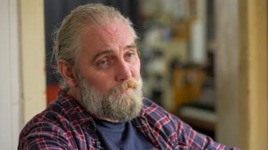 Adam, a man with long hair and a beard, wearing a chequered shirt, looks at the camera with a neutral expression