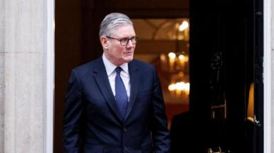 Prime Minister Sir Keir Starmer standing in front of an open door to 10 Downing Street in London, in a navy suit and blue tie looking to the right.