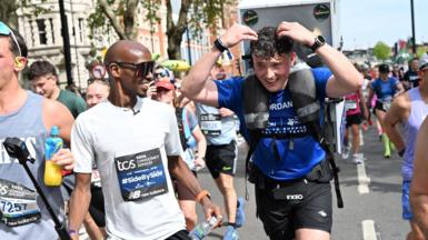 Jordan Adams wearing a blue t-shirt and black running shorts runs with a white fridge on his back surrounded by other runners. Mo Farah, wearing a white t-shirt and sunglasses, runs beside him.