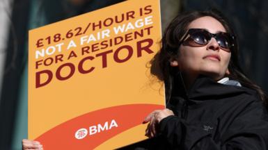 A woman with brown hair wearing sunglasses holds an orange BMA branded placard which reads: "£18.62/hour is not a fair wage for a resident doctor"