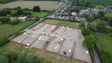 A drone image of the developed site. It is a field that has been largely concreted over and has several caravans, both static and mobile, on it. Fencing is being installed around the perimeter of the site, which is flanked by residential housing and other fields.