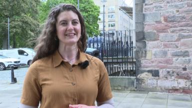 Boireannach le falt fada donn na seasamh air sràid ann an Dun Eideann, le gàire air a h-aodann. Tha leìne dorch buidhe oirre.