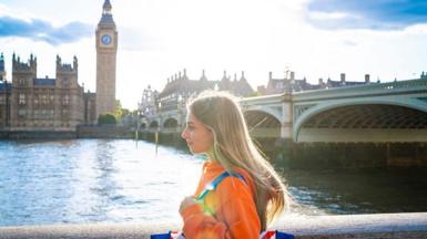 A young woman wearing a bright orange jacket and a Union Jack-coloured bag on her shoulder walks along the bank of the river Thames. Big Ben and the Houses of Parliament is in the background behind her.