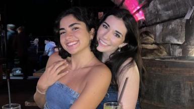 Two girls smiling at the camera. On the left, the girl has shoulder-length dark hair. She is wearing a blue denim top and silver necklace. The girl on the right has long dark hair. She is wearing a blue denim top and silver earrings. The photo is taken in a bar with long wooden tables in the background.