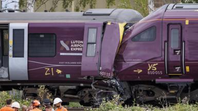 Two damaged trains, the front of one purple train is crushed into the end of another, with severe impact damage visible around the cab area.