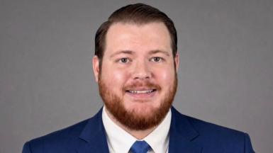 A man with short, coiffed hair, and a short ginger beard is smiling at the camera. He is wearing a blue suit and tie.