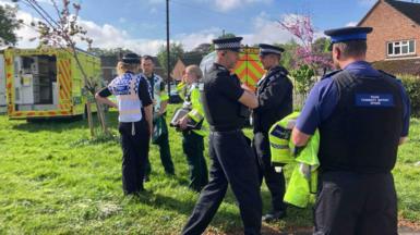 PCSOs, police officers and paramedics are standing on an area of grass in a residential area chatting. 